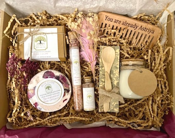 May include: A gift box containing a bar of Rose Glow soap, a round bath bomb with dried rose petals, a glass tube of bath salts, a small glass bottle of bath oil, a wooden spoon, a candle in a glass jar, and a wooden comb engraved with the text You are already Astriya.