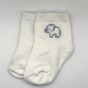 Könnte beinhalten: Ein Paar weiße Söckchen mit einer kleinen, aufgestickten blauen Elefantenkontur an der Außenseite jeder Socke. Die Socken haben ein geripptes Bündchen am oberen Rand und eine schlichte, einfarbig weiße Strickstruktur im Fuß- und Fersenbereich.