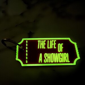May include: A rectangular keychain with rounded edges and a metal ring. The keychain has a dark red background with the words "THE LIFE OF A SHOWGIRL" in bright green. The edges of the keychain are also green.