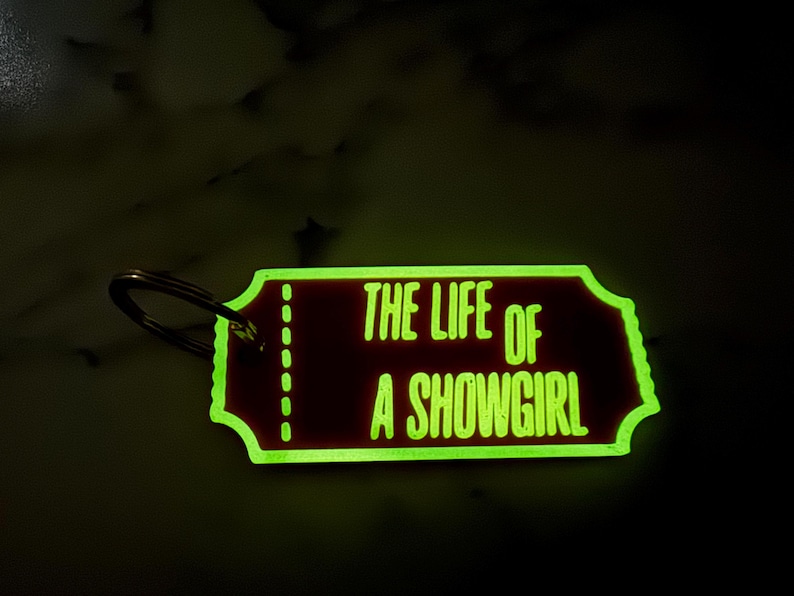 May include: A rectangular keychain with rounded edges and a metal ring. The keychain has a dark red background with the words "THE LIFE OF A SHOWGIRL" in bright green. The edges of the keychain are also green.