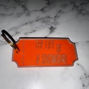 May include: An orange keychain with the text "THE LIFE OF A SHOWGIRL" in a serif font. The keychain has a ticket shape with a gold key ring attached.