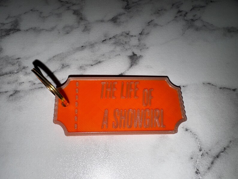 May include: An orange keychain with the text "THE LIFE OF A SHOWGIRL" in a serif font. The keychain has a ticket shape with a gold key ring attached.