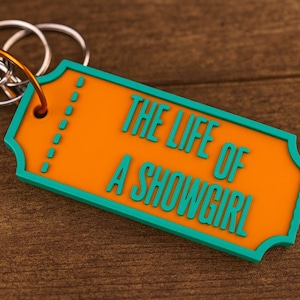 May include: An orange and teal keychain with the text "THE LIFE OF A SHOWGIRL". The keychain is shaped like a ticket and has a teal border. The text is in teal and is in a bold, sans-serif font. The keychain is attached to a silver keyring.
