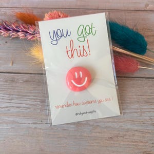 May include: A round coral-colored smiley face pin attached to a white display card. The card features the text you got this! in blue and green, and remember how awesome you are! in red, with the social media handle @rubyandrosegifts at the bottom.