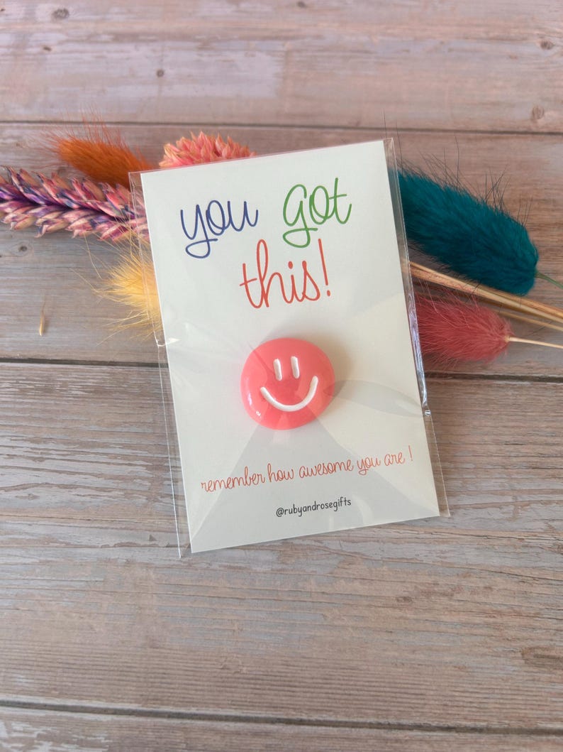 May include: A round coral-colored smiley face pin attached to a white display card. The card features the text you got this! in blue and green, and remember how awesome you are! in red, with the social media handle @rubyandrosegifts at the bottom.
