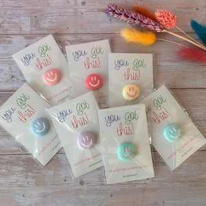 May include: Seven individual circular pins, each featuring a different pastel-colored smiley face, are attached to white display cards. Each card displays the text "you got this!" in colorful script and is individually wrapped in clear plastic.