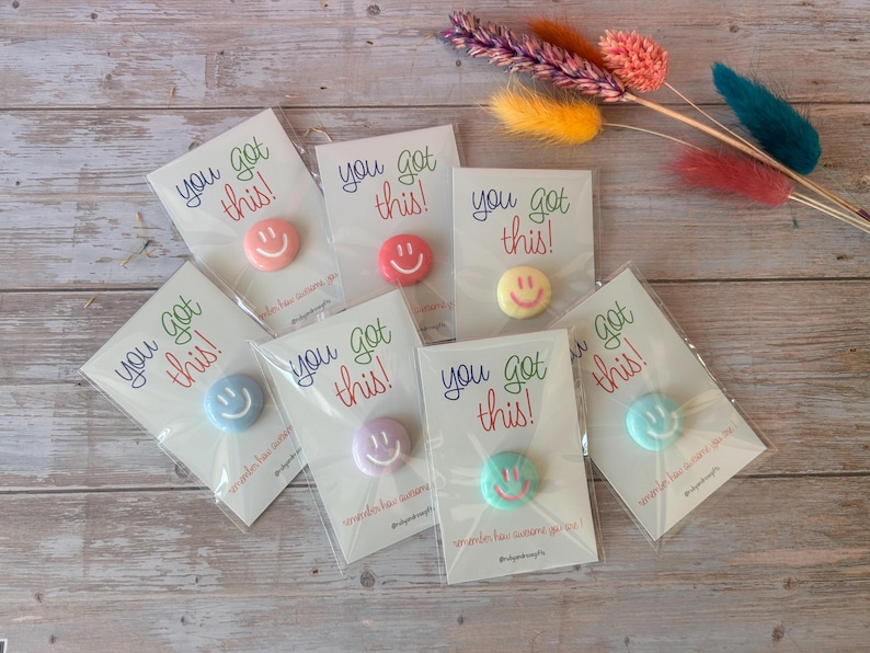 May include: Seven individual circular pins, each featuring a different pastel-colored smiley face, are attached to white display cards. Each card displays the text "you got this!" in colorful script and is individually wrapped in clear plastic.