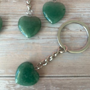 May include: A collection of heart-shaped keyrings made from polished green gemstones. Each heart is connected to a silver-coloured chain and keyring. The keyrings are displayed on a light wooden surface.