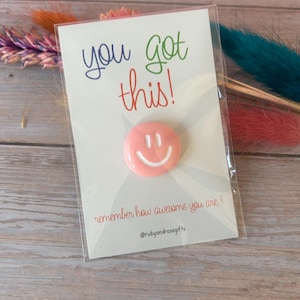 May include: A circular pink pin with a white smiley face graphic attached to a white card. The card features the text you got this! in blue and green, the phrase remember how awesome you are! in red, and the handle @rubyandrosegifts at the bottom.