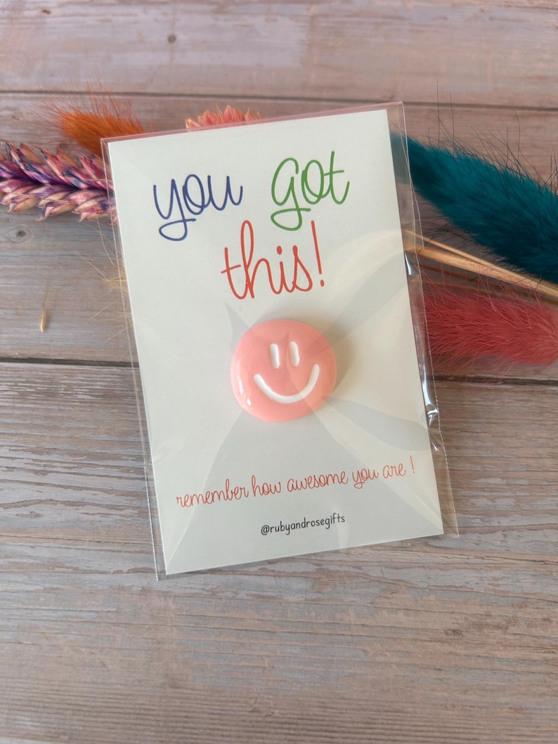 May include: A circular pink pin with a white smiley face graphic attached to a white card. The card features the text you got this! in blue and green, the phrase remember how awesome you are! in red, and the handle @rubyandrosegifts at the bottom.