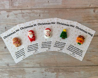May include: Five white display cards featuring Christmas-themed clay charms: a gingerbread person, Santa Claus, a snowman, a Christmas tree, and a reindeer. Each card includes the text Christmas Hug and a poem about sending holiday cheer.