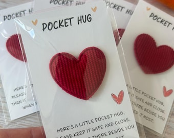 May include: A red fabric heart token attached to a white card inside a clear plastic sleeve. The card features the text POCKET HUG and a poem that reads: HERE'S A LITTLE POCKET HUG, PLEASE KEEP IT SAFE AND CLOSE. THEN IT WILL BE THERE BESIDE YOU WHEN YOU NEED IT MOST.