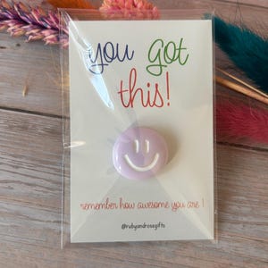 May include: A light purple circular pin with a white smiley face design, attached to a white card featuring the text "you got this!" in blue, green, and red, and "remember how awesome you are !" in red, with the handle @rubyandrosegifts at the bottom.
