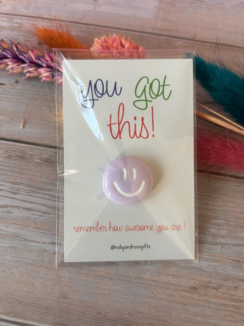 May include: A light purple circular pin with a white smiley face design, attached to a white card featuring the text "you got this!" in blue, green, and red, and "remember how awesome you are !" in red, with the handle @rubyandrosegifts at the bottom.