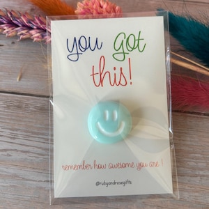May include: A light blue circular smiley face pin attached to a white card in a clear plastic sleeve. The card features the text you got this! in blue, green, and red, with the text remember how awesome you are! and @rubyandrosegifts below.