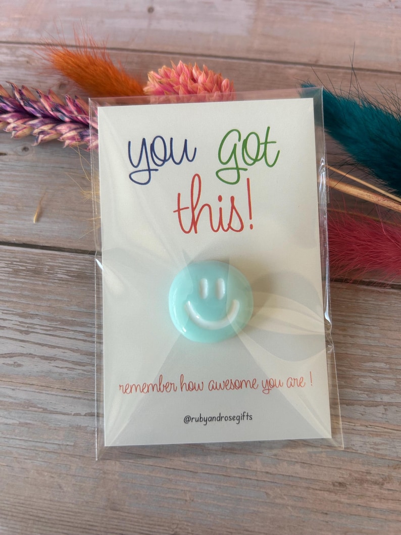 May include: A light blue circular smiley face pin attached to a white card in a clear plastic sleeve. The card features the text you got this! in blue, green, and red, with the text remember how awesome you are! and @rubyandrosegifts below.