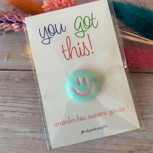 May include: A round mint green pin with a pink smiley face design attached to a white card. The card features the text "you got this!" in blue and red, "remember how awesome you are !" in red, and the social media handle "@rubyandrosegifts".