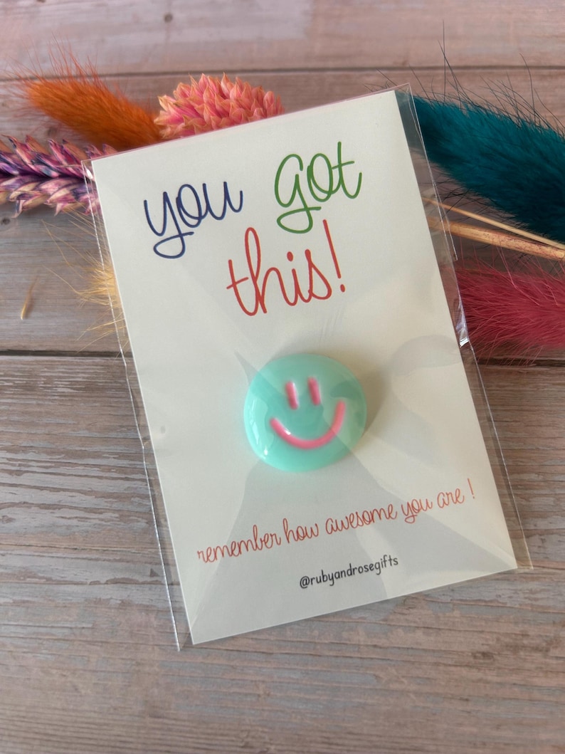 May include: A round mint green pin with a pink smiley face design attached to a white card. The card features the text "you got this!" in blue and red, "remember how awesome you are !" in red, and the social media handle "@rubyandrosegifts".