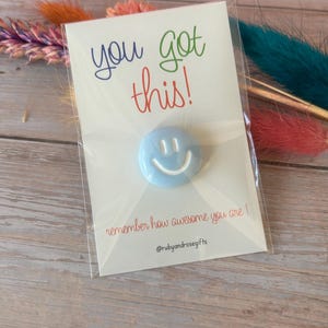 May include: A light blue circular pin with a white smiley face graphic attached to a white card. The card features the text "you got this!" in blue, green, and red, with "remember how awesome you are!" and "@rubyandrosegifts" printed below.