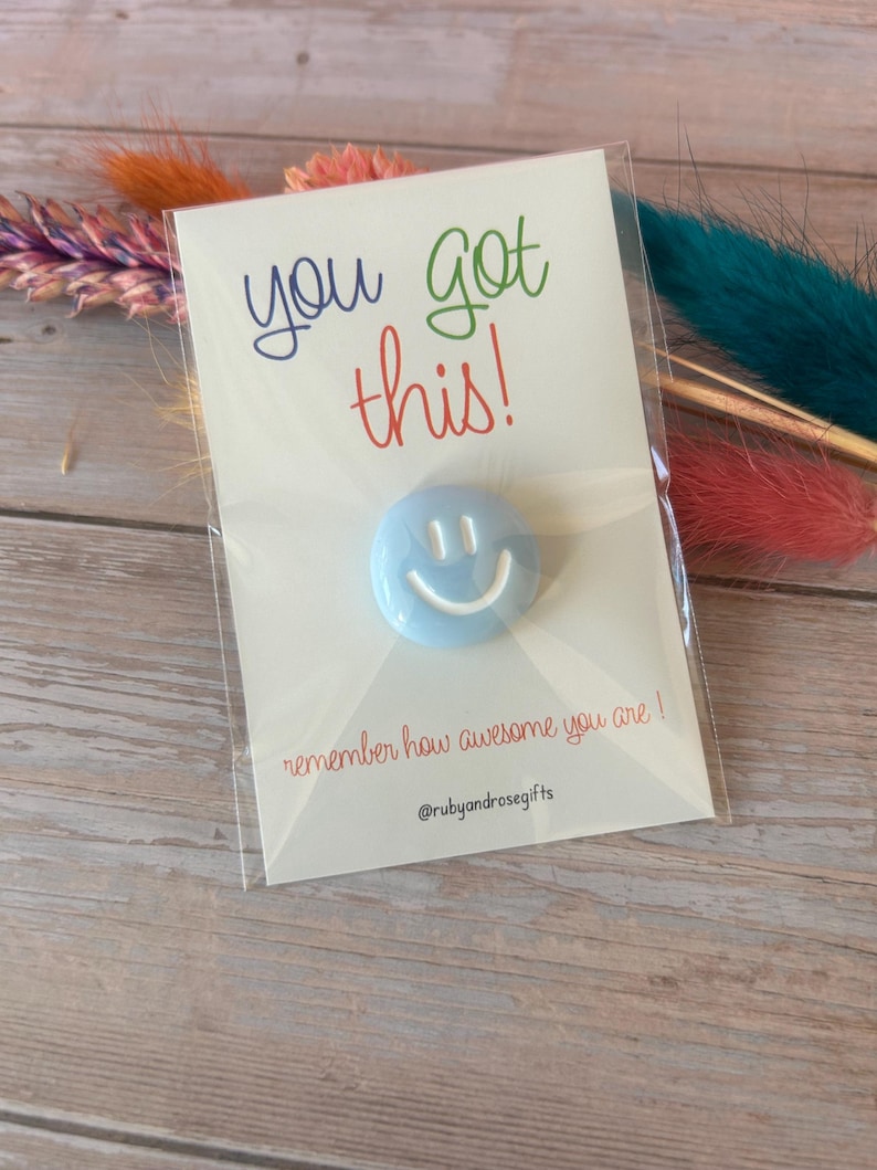 May include: A light blue circular pin with a white smiley face graphic attached to a white card. The card features the text "you got this!" in blue, green, and red, with "remember how awesome you are!" and "@rubyandrosegifts" printed below.