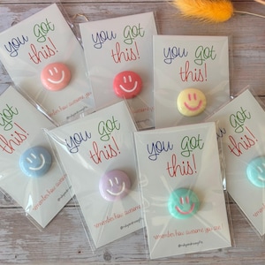 May include: Seven circular pins in various pastel colors, each featuring a white smiley face. Each pin is attached to a white card printed with the text you got this! and remember how awesome you are! in blue, green, and red lettering.