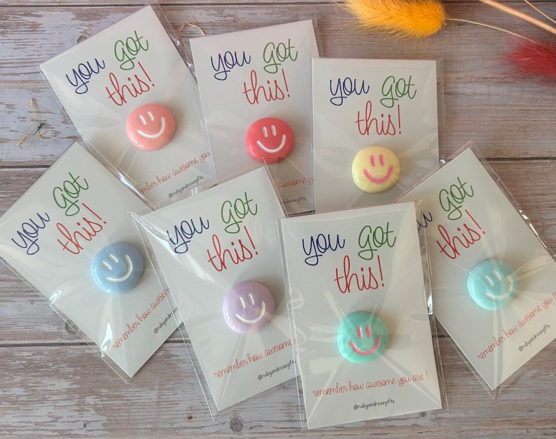 May include: Seven circular pins in various pastel colors, each featuring a white smiley face. Each pin is attached to a white card printed with the text you got this! and remember how awesome you are! in blue, green, and red lettering.