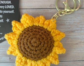May include: A crochet sunflower keychain featuring a dark brown center and bright yellow petals. The flower is attached to a gold-toned metal split ring and lobster clasp. A black card with white text reading ENOUGH, very LOVED, to yourself, ay, elf is visible.