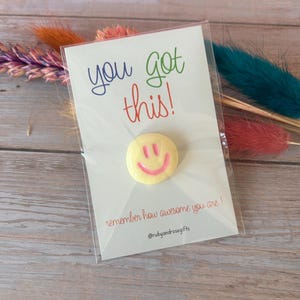 May include: A circular yellow pin with a pink smiley face design attached to a white display card. The card features the text you got this! in blue and green, and the text remember how awesome you are! and @rubyandrosegifts in red.