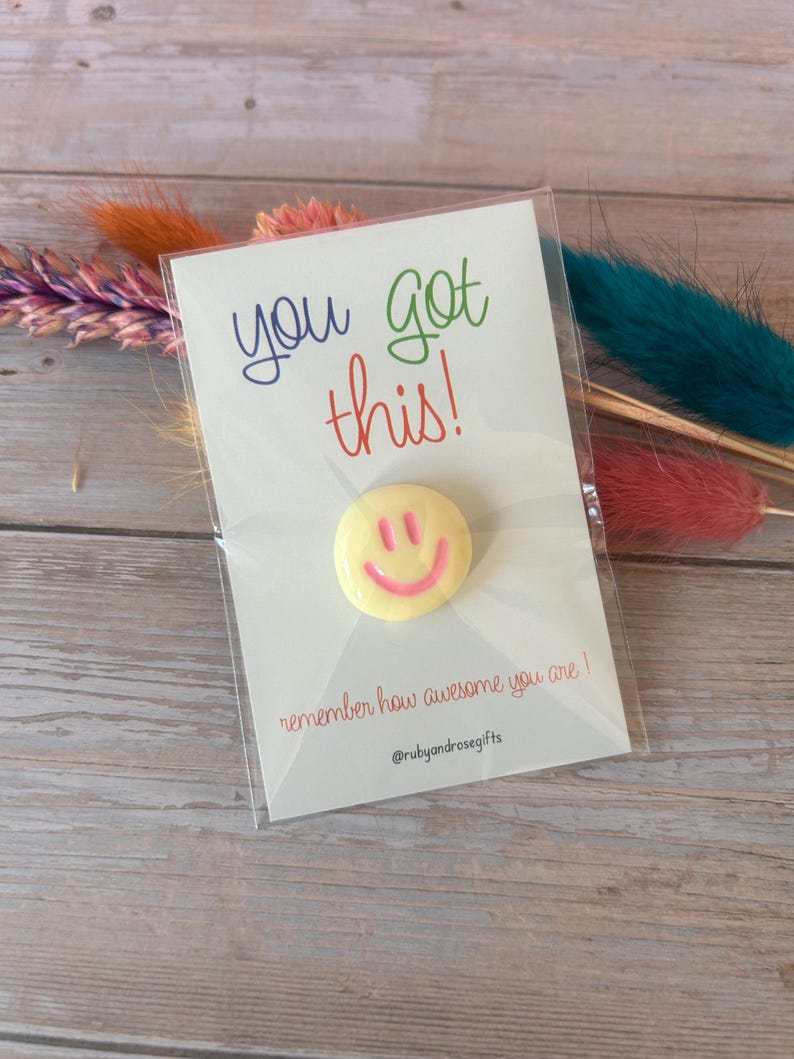 May include: A circular yellow pin with a pink smiley face design attached to a white display card. The card features the text you got this! in blue and green, and the text remember how awesome you are! and @rubyandrosegifts in red.