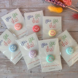 May include: Seven individual circular pins in various pastel colours, each featuring a smiling face. Each pin is attached to a white card printed with the text you got this! in colourful script, packaged in a clear plastic sleeve for protection.