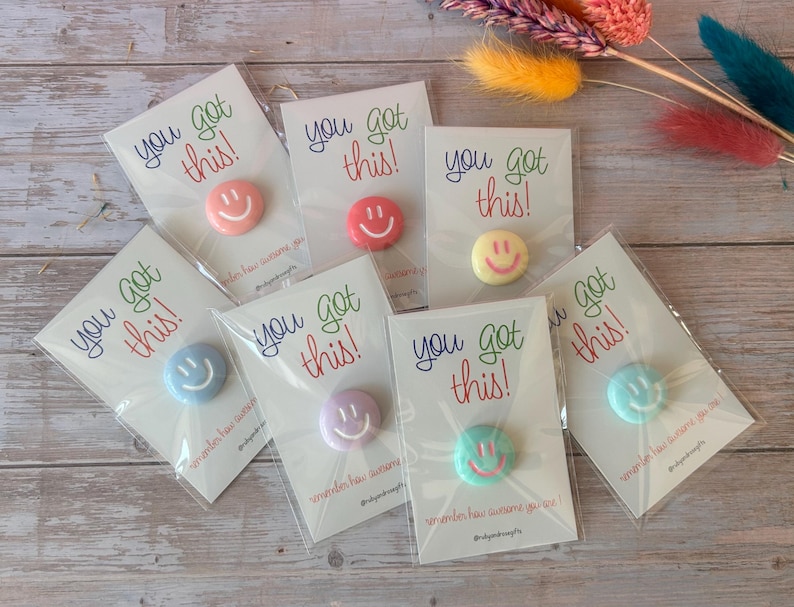 May include: Seven individual circular pins in various pastel colors, each featuring a smiling face. Each pin is attached to a white card printed with the text you got this! in colorful script, packaged in a clear plastic sleeve for protection.