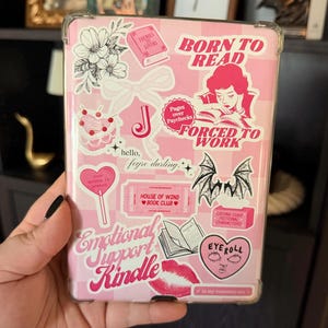 May include: Pink Kindle case with a checkered pattern and various stickers. Stickers include floral designs, a bow, a cake, and text that reads "BORN TO READ", "FORCED TO WORK", and "Emotional Support Kindle".