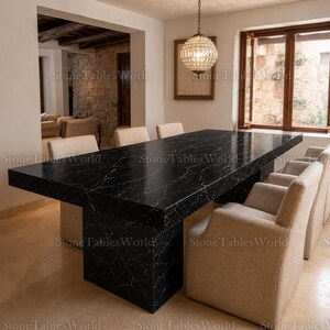 May include: A large, rectangular black marble dining table with white veining. The table is surrounded by beige upholstered chairs. A decorative chandelier hangs above the table, and a window with wooden frames is visible in the background.