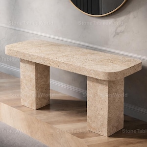 以下が含まれることがあります： 長方形の天板と2つの正方形の脚を備えたベージュ色の石のコンソールテーブル。テーブルは丸みを帯びたエッジと斑点のあるテクスチャを持っています。テーブルは木製の床に置かれています。