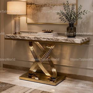 Può includere: Una consolle con piano in marmo e base dorata a gambe incrociate. Il tavolo ha una forma rettangolare con finiture dorate. Una lampada con paralume beige e base decorativa è appoggiata sul tavolo. La stanza è ben illuminata.