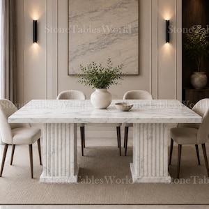 Puede incluir: Una mesa de comedor rectangular de mármol blanco con superficie pulida y dos pedestales estriados en forma de columna. La mesa está rodeada por cuatro sillas tapizadas con patas de madera oscura y un jarrón blanco con follaje verde.