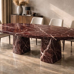 May include: An oval dining table with a burgundy marble top and two pedestal bases. The table is surrounded by four cream-coloured chairs with wooden legs. The room has a neutral colour palette with natural light.