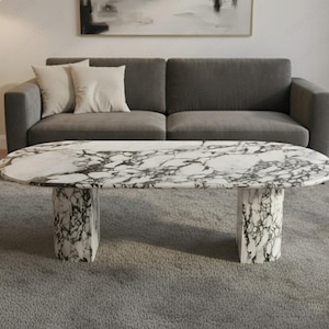 Puede incluir: Mesa de centro ovalada de mármol con una parte superior con estampado blanco y negro. La mesa tiene dos bases cilíndricas. La mesa está frente a un sofá gris con dos cojines color crema. La habitación tiene una alfombra gris.