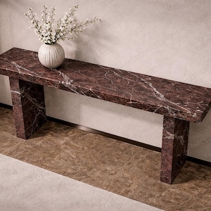 May include: A rectangular marble console table with a dark red and white veined pattern. The table has two rectangular legs and a white vase with white flowers on top. The table is 152 cm long.