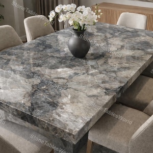 May include: A large rectangular dining table with a gray and white patterned stone top. The table is surrounded by eight beige upholstered chairs. A black vase with white flowers sits in the center of the table. The table is approximately 100 inches long.