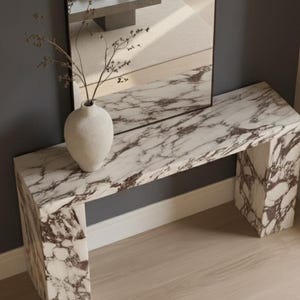 Puede incluir: Una consola de mármol con un patrón veteado blanco y marrón. Un jarrón blanco con ramas secas se encuentra sobre la mesa. Un espejo está montado en la pared detrás de la mesa.