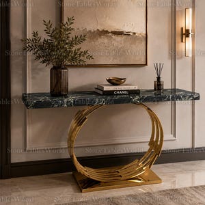 Può includere: Una consolle moderna con piano in marmo nero e verde e base circolare dorata. Il tavolo è decorato con un vaso di verde, libri e una ciotola decorativa. Il tavolo è lungo circa 122 cm.