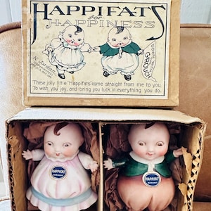 以下が含まれることがあります： ヴィンテージのHappifat人形のオリジナルボックス。ボックスには「Happifats for Happiness」を含むイラストとテキストが描かれています。人形は段ボール箱に入っており、1つはピンクのドレス、もう1つはグリーンのジャケットを着ています。各人形には「Happifat Germany」のラベルが付いています。