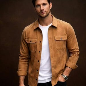 May include: A tan suede shirt jacket with two chest pockets and dark brown buttons. The jacket is worn over a white t-shirt. The person is wearing a silver watch and black pants. The background is a solid brown.