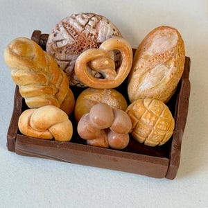 Può includere: Un vassoio di legno marrone pieno di prodotti da forno in miniatura. L'assortimento comprende un pretzel, una baguette, un pane rotondo e altri tipi di pane. Il pane è in varie tonalità di marrone e beige.