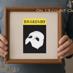 May include: A framed needlepoint cross stitch chart featuring a black and white design of a Phantom of the Opera mask. The word "BROADWAY" is in yellow letters above the mask. The framed artwork is 10 cm x 15 cm on 13-count canvas.