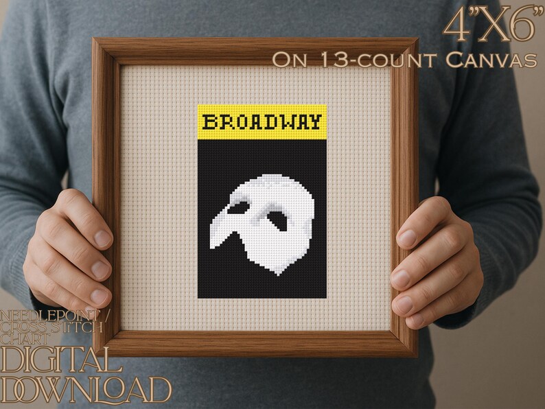 May include: A framed needlepoint cross stitch chart featuring a black and white design of a Phantom of the Opera mask. The word "BROADWAY" is in yellow letters above the mask. The framed artwork is 10 cm x 15 cm on 13-count canvas.
