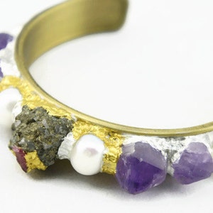 Pode incluir: Uma pulseira de punho dourada com uma faixa prateada e dourada. A pulseira é adornada com pedras de ametista roxas, pérolas brancas e um aglomerado de minerais cinza escuro e vermelhos. Uma peça de joalharia única.