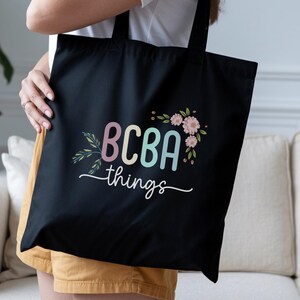 May include: Black tote bag featuring the text "BCBA things" in pastel shades, adorned with floral details. The bag is held by a person wearing a white top and yellow shorts. A stylish and practical accessory.