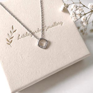 May include: A silver-toned necklace featuring a small, rounded square pendant with embedded clear stones. The necklace rests on a cream-coloured box with the text Little Garden Jewellery and a gold leaf illustration printed on the surface.